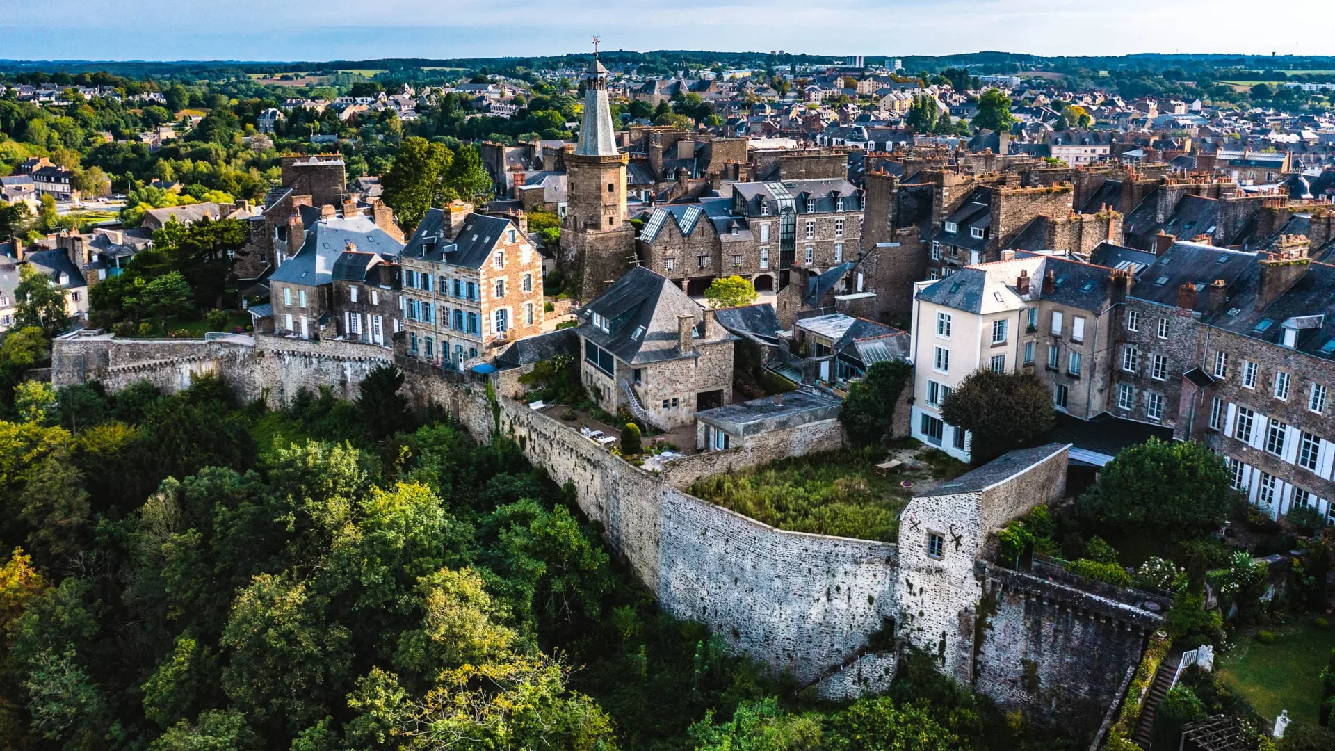 Ville médiévale avec maisons en pierre et toit en ardoises avec de nombreux arbres implanté dans la ville.